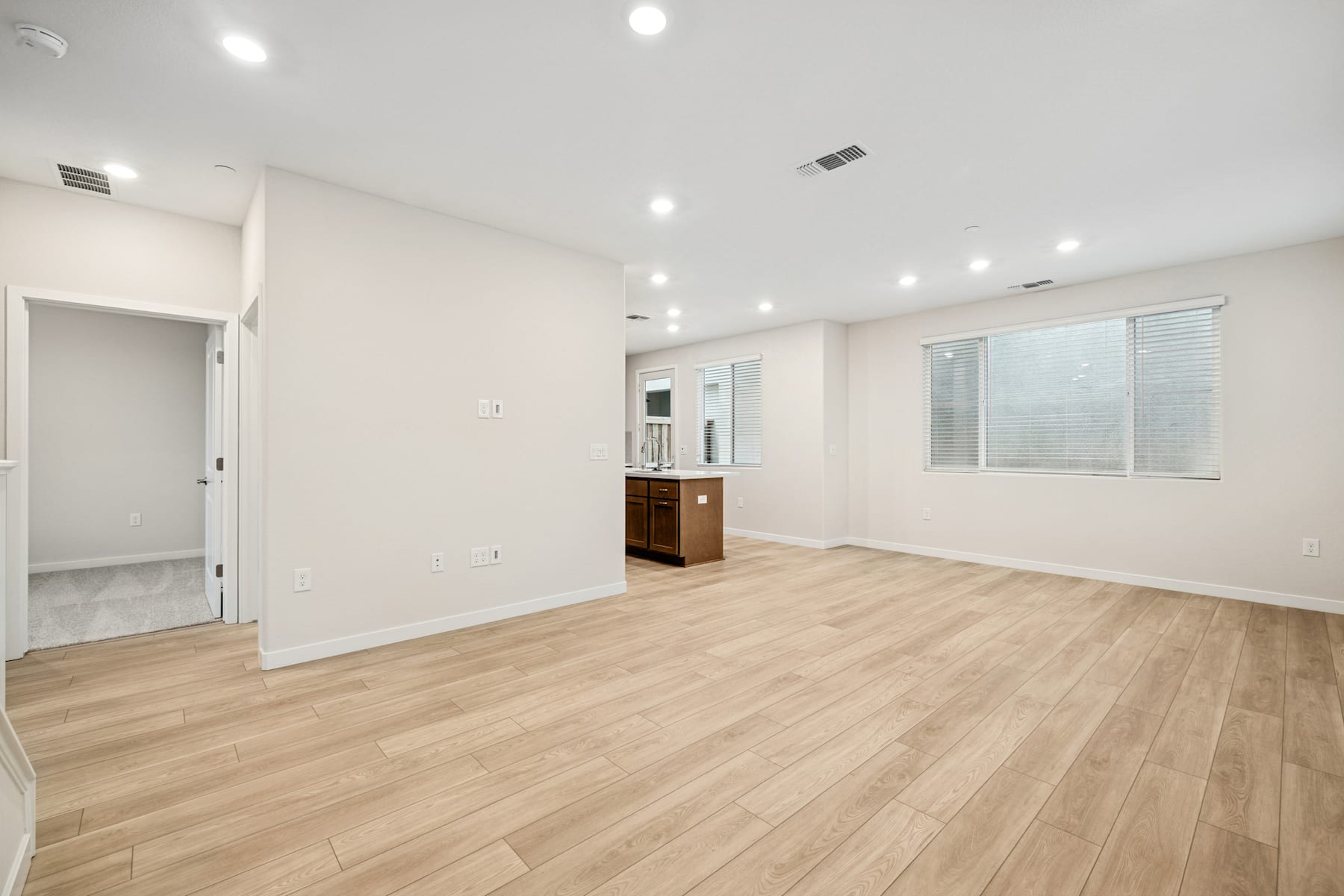 A spacious, well-lit room with light-colored hardwood floors, white walls, and recessed lighting fixtures. A wooden cabinet or dresser is visible in the background.