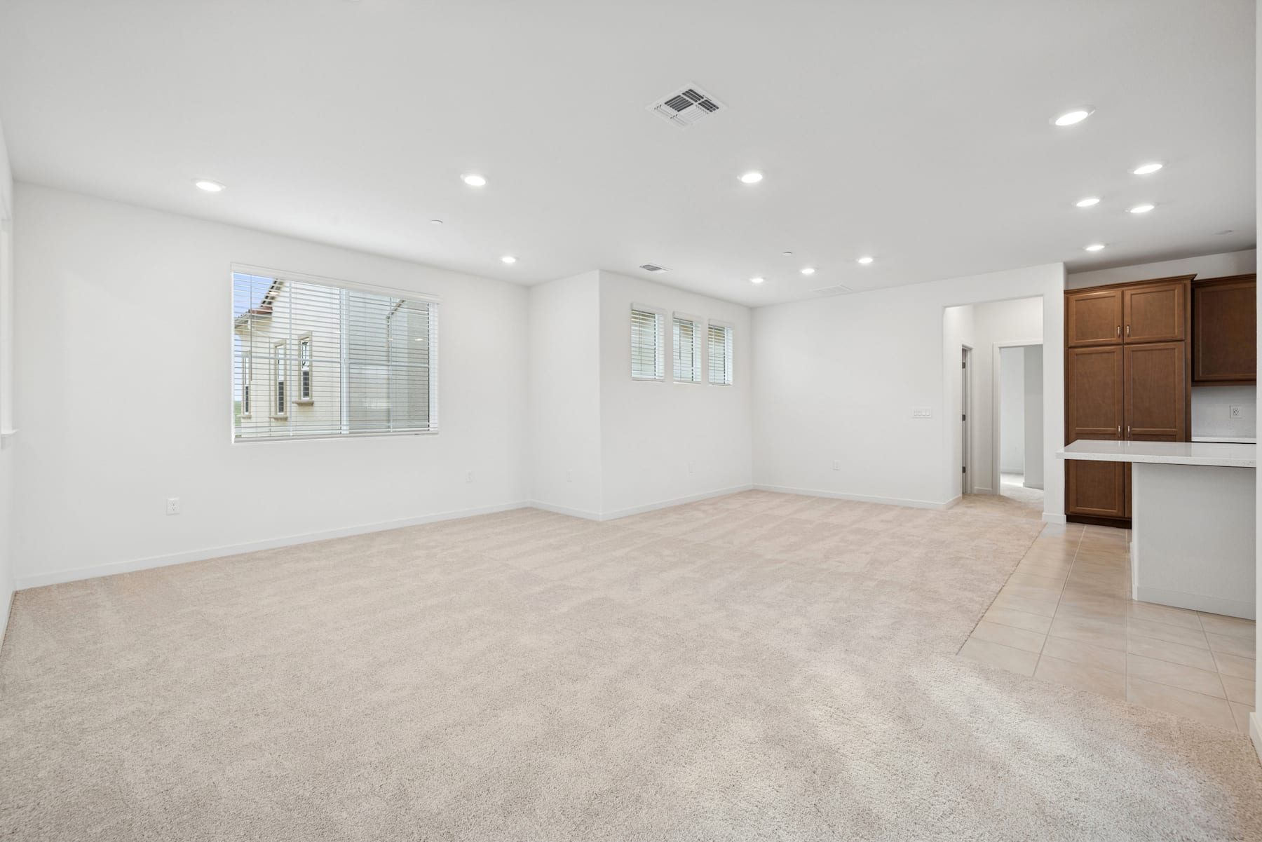 A spacious, well-lit room with a clean, minimalist design featuring a large window, recessed lighting, and wooden cabinets in the background.