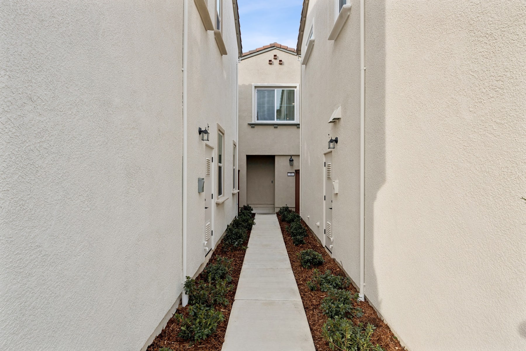 A narrow walkway lined with potted plants leads to a white building with a decorative arch at the end.