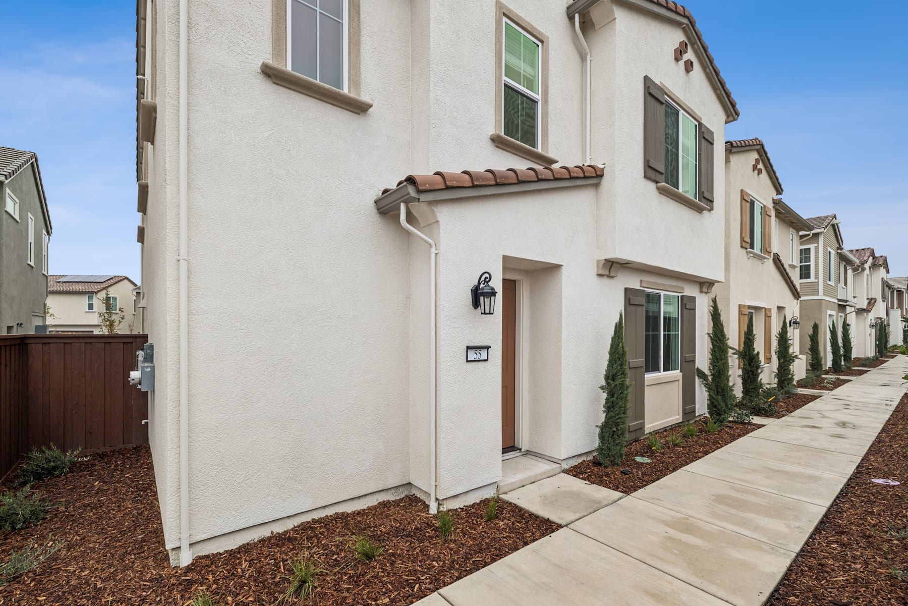 A row of modern, two-story townhouses with beige stucco exteriors, tiled roofs, and landscaped yards in the foreground, set against a clear blue sky in the background.