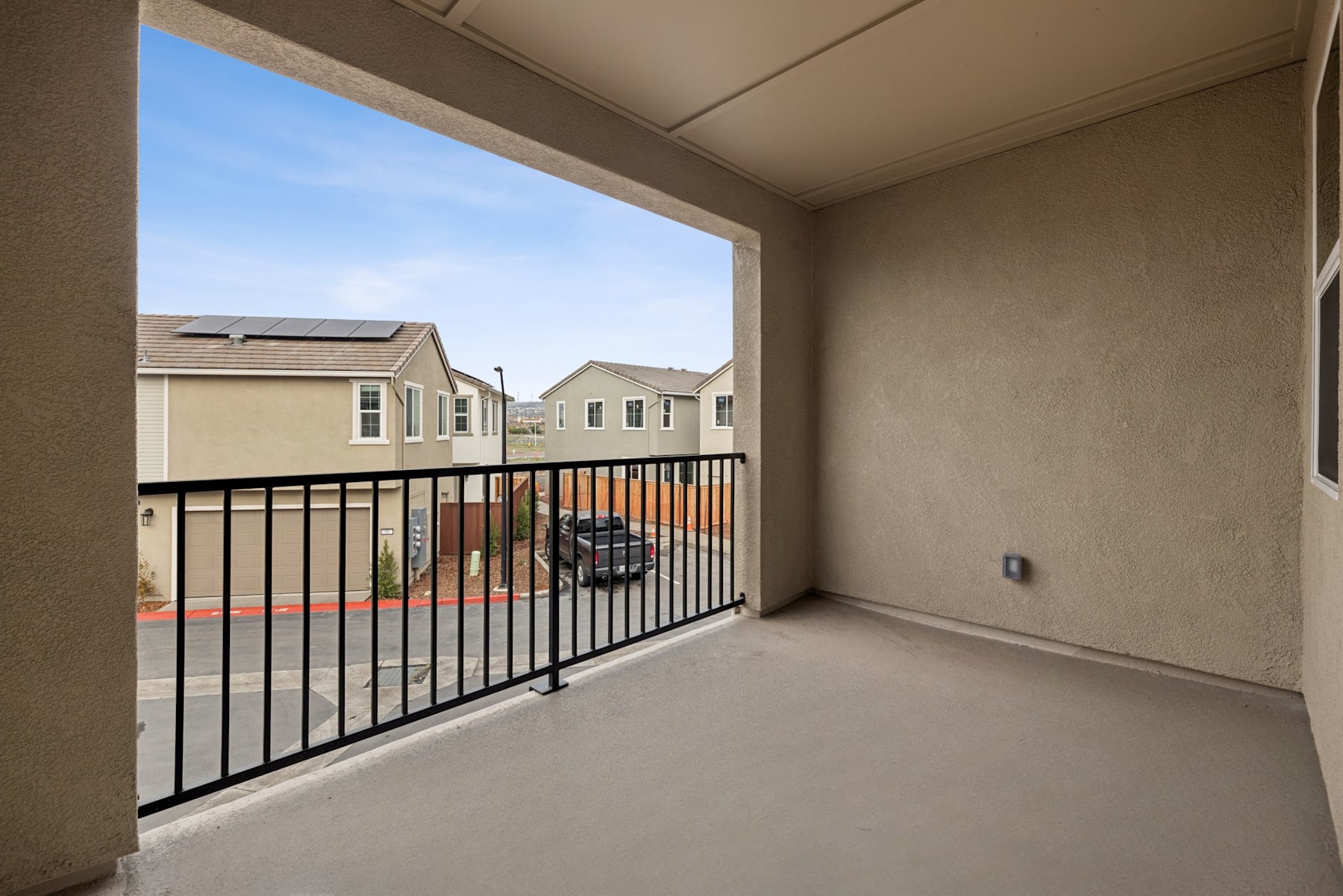 A balcony with a metal railing overlooks a residential neighborhood with several houses in the background under a clear blue sky.
