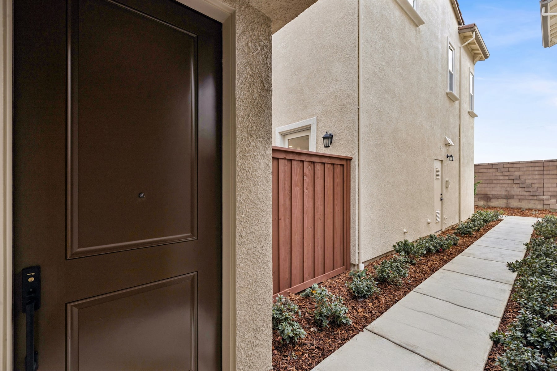A brown wooden door leads to a walkway lined with shrubs and a concrete path, with a beige stucco building visible in the background.