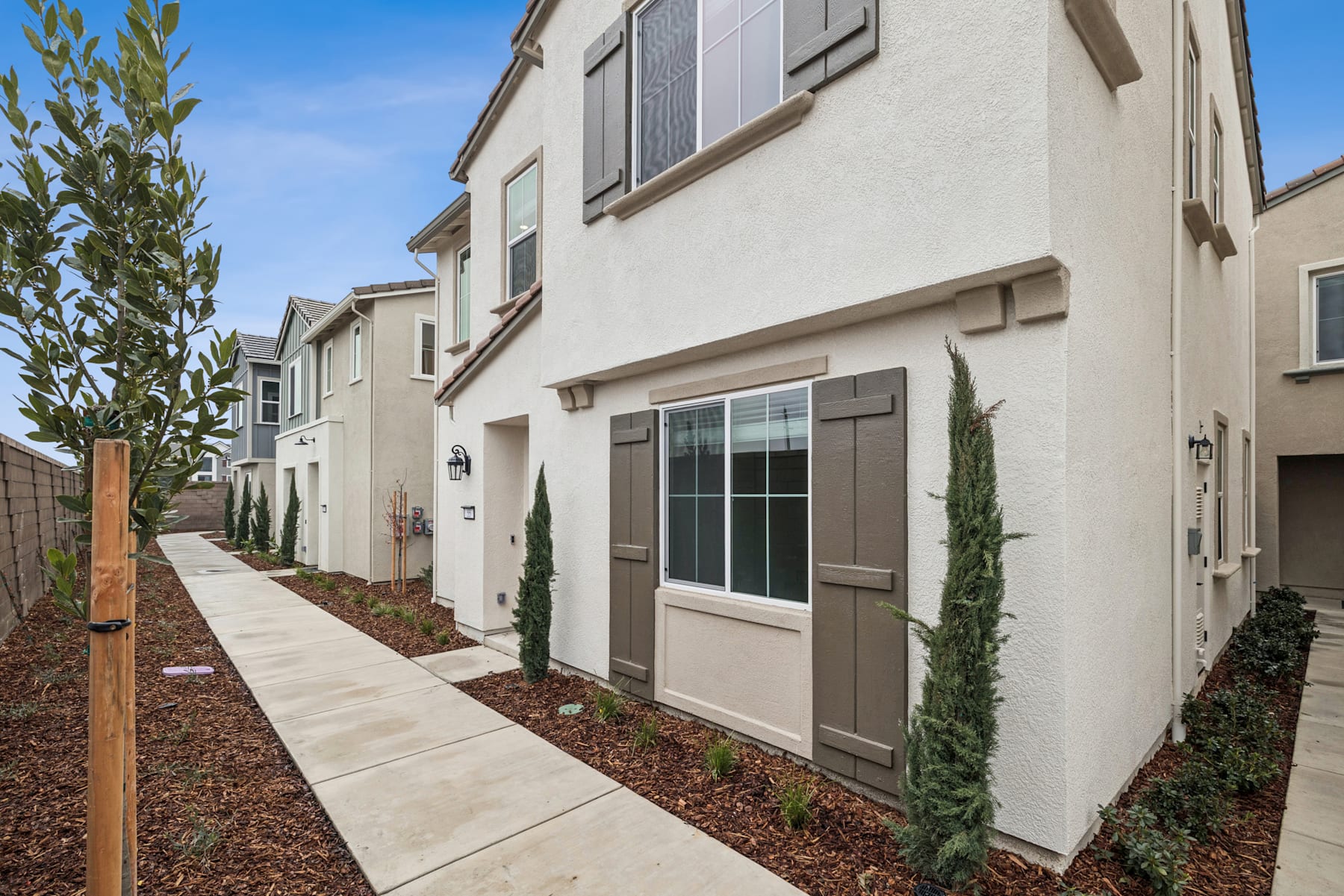 A row of newly constructed townhouses with a paved walkway and landscaping in the foreground, set against a clear blue sky.