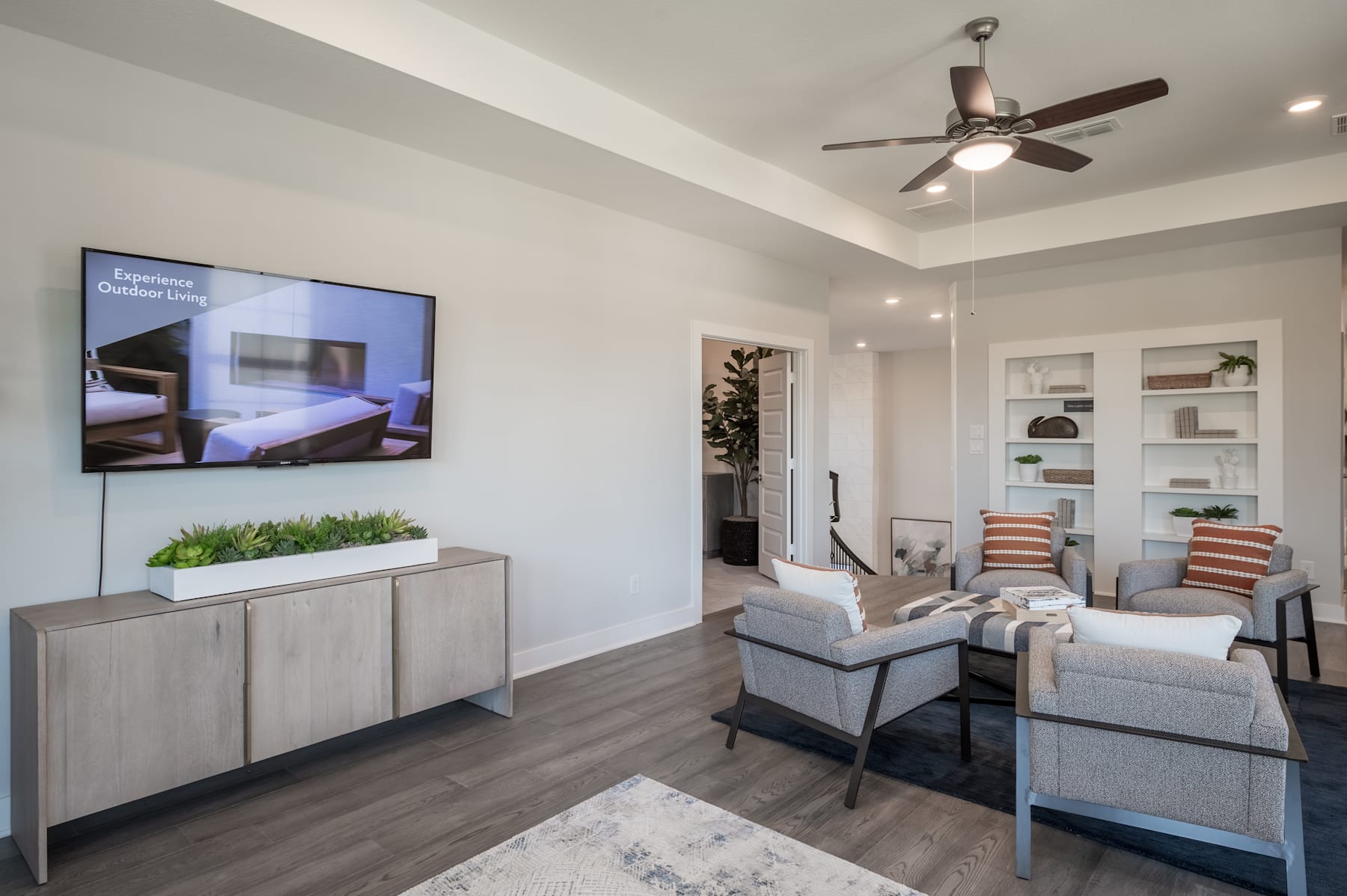 A modern and cozy living room with a large TV, comfortable seating, and a ceiling fan, set against a neutral color palette and wooden flooring.