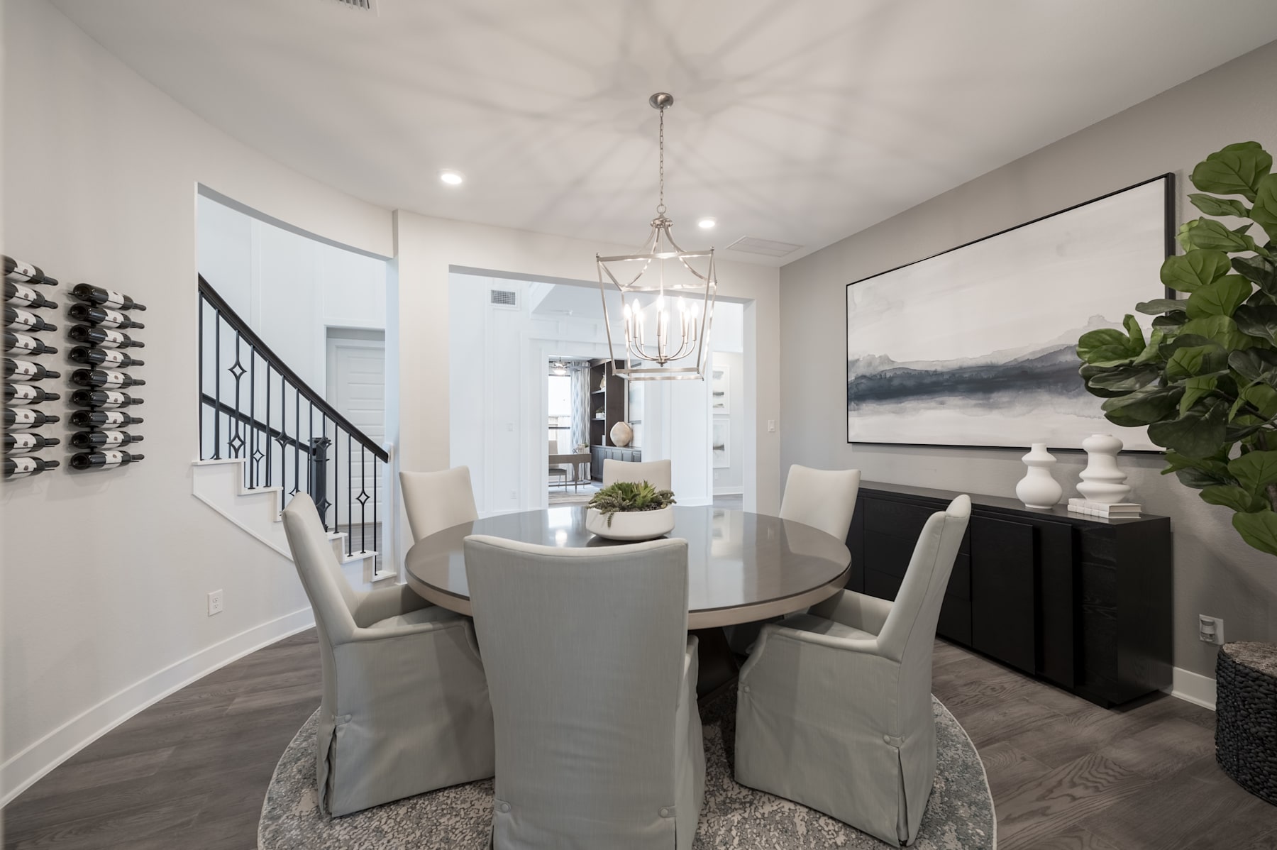 A modern and stylish dining room with a round table, upholstered chairs, and a chandelier, set against a backdrop of a wall art piece and a wine rack.