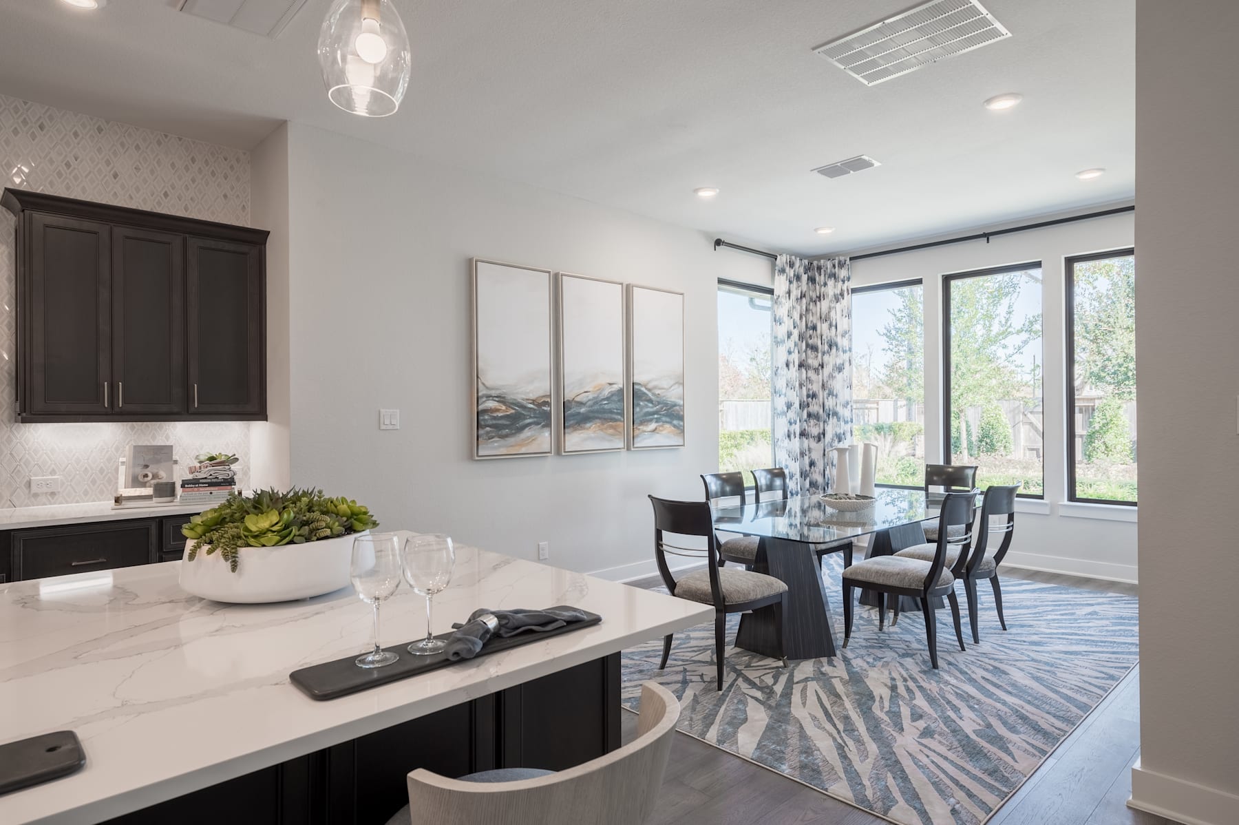 A modern and stylish dining room with a sleek kitchen counter, glass-paneled cabinets, and a dining table set with chairs, all surrounded by large windows overlooking a scenic outdoor view.