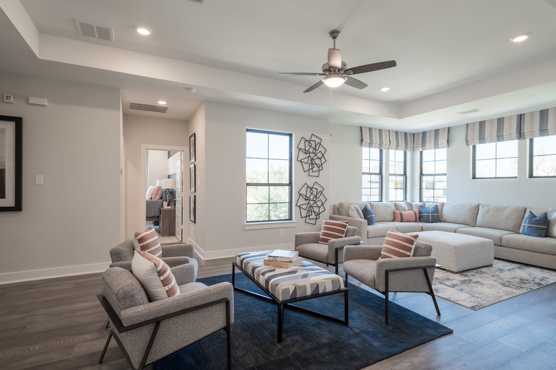 A spacious and well-furnished living room with a large sectional sofa, armchairs, and a patterned rug, complemented by a ceiling fan and large windows providing natural light.