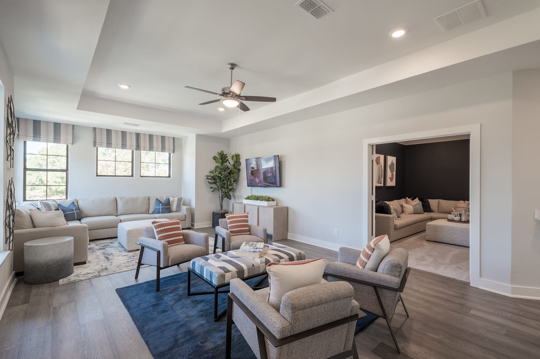 A spacious and well-furnished living room with comfortable seating arrangements, a ceiling fan, and large windows providing natural light, set against a backdrop of a cozy and inviting atmosphere.