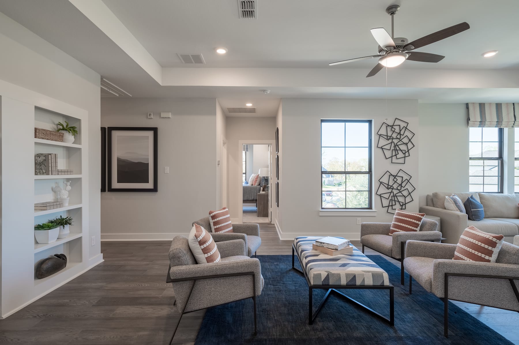 A modern and cozy living room with comfortable gray sofas, a blue patterned rug, and decorative wall art, complemented by a ceiling fan and shelving unit in the background.