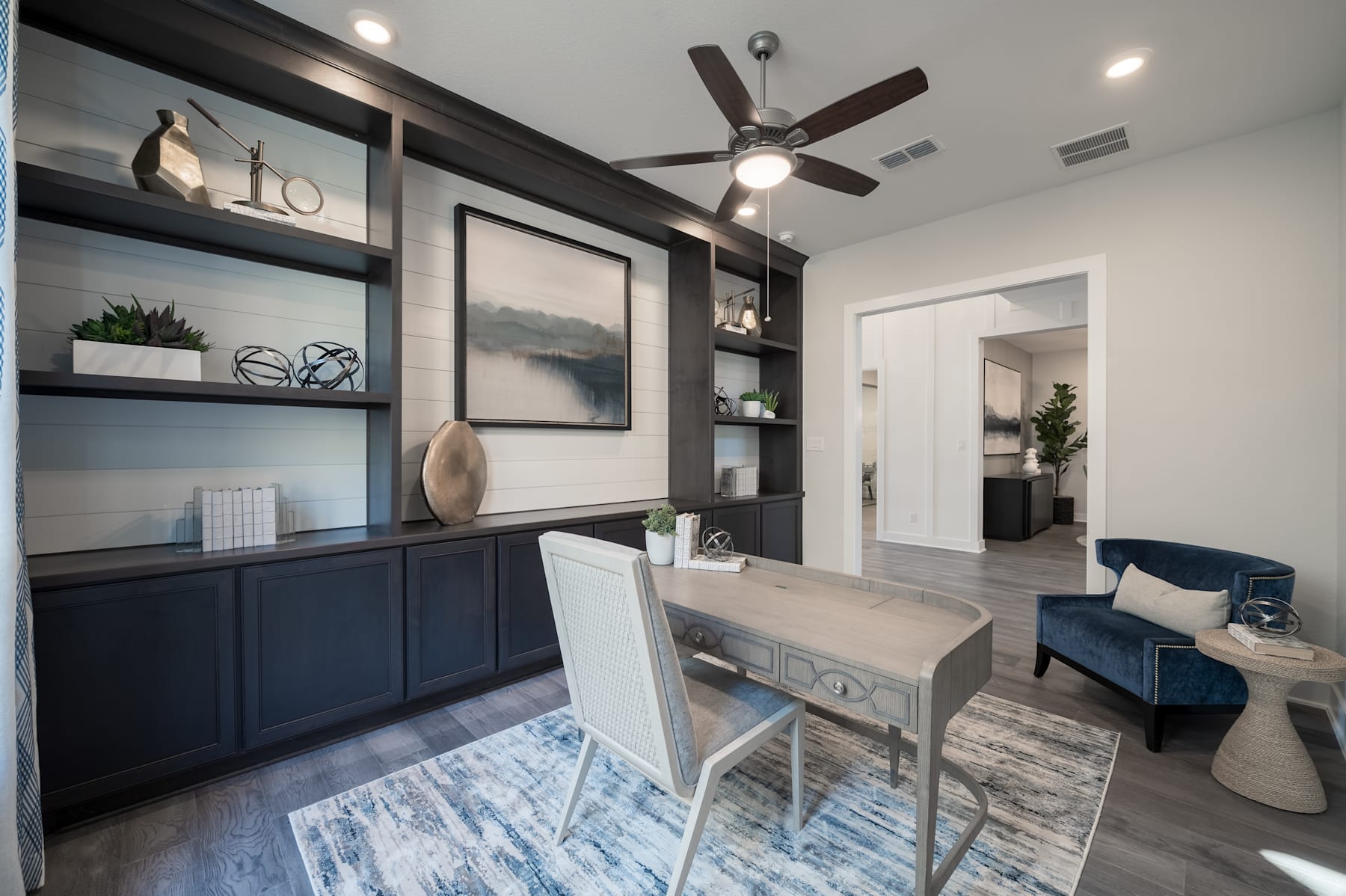 A modern and stylish home office space with built-in shelving, a desk, and a ceiling fan, set against a neutral color palette and accented with potted plants and artwork.