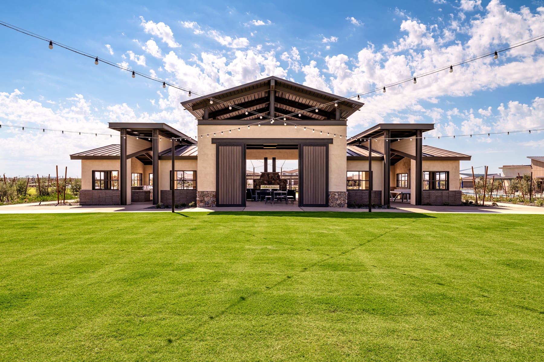 A large, modern building with a wooden exterior stands in the foreground, surrounded by a lush green lawn. The sky in the background is filled with fluffy white clouds against a bright blue backdrop.