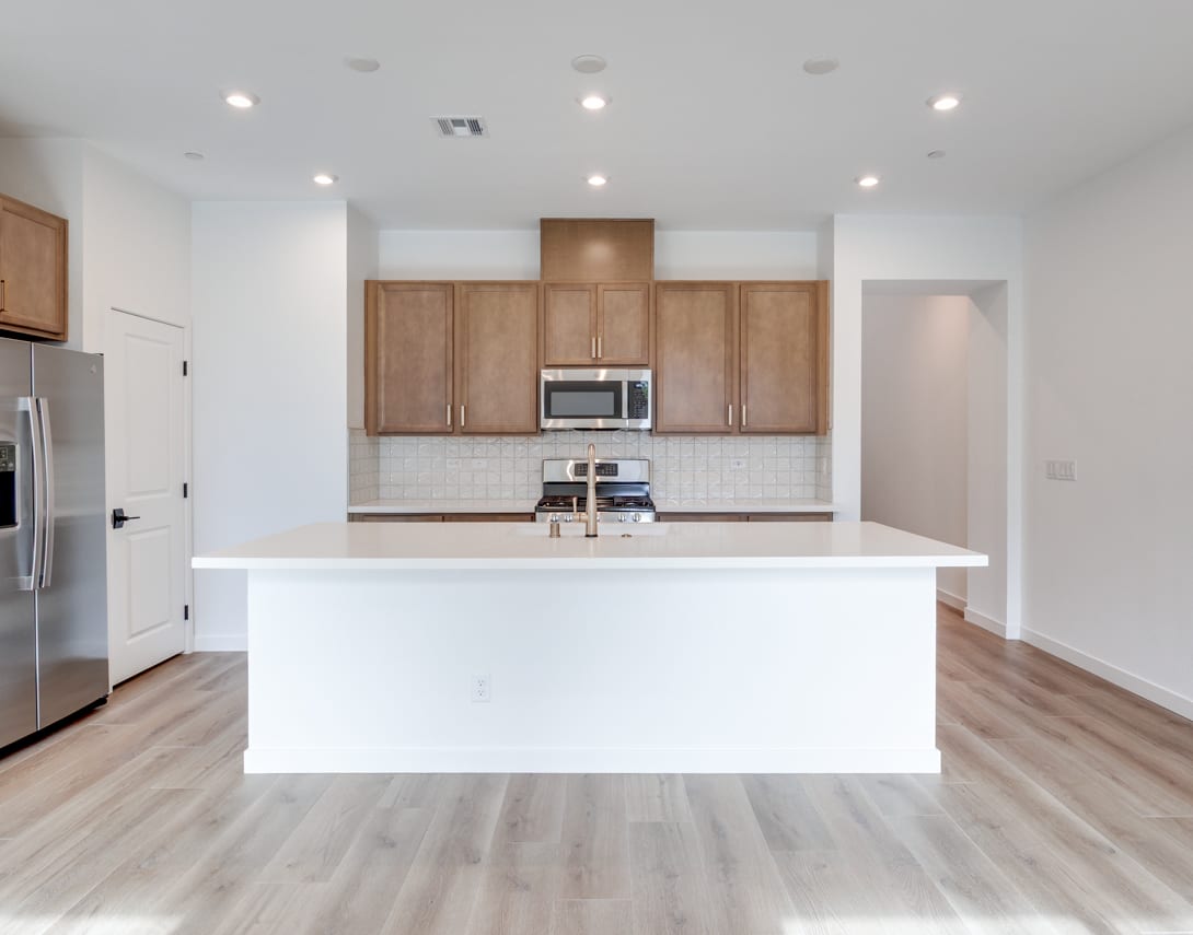 A modern, open-concept kitchen with white countertops, wooden cabinets, and stainless steel appliances, set against a backdrop of hardwood floors and recessed lighting.