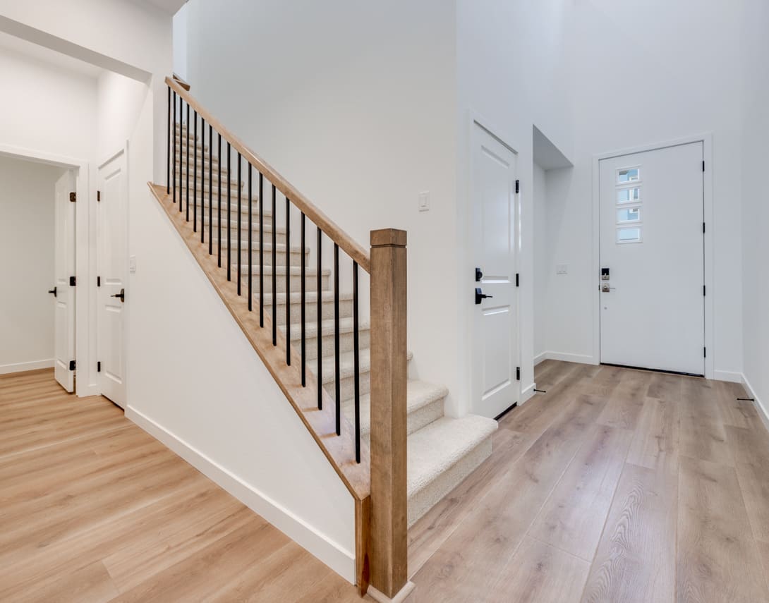 A bright and spacious entryway with a wooden staircase and railing, leading to a hallway with a white door and hardwood flooring.