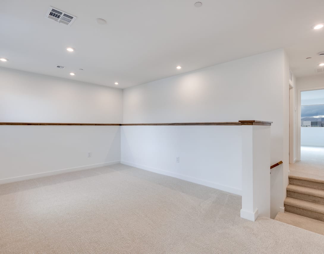 A spacious, well-lit room with white walls, a wooden handrail, and a carpeted floor, suggesting a modern and minimalist interior design.