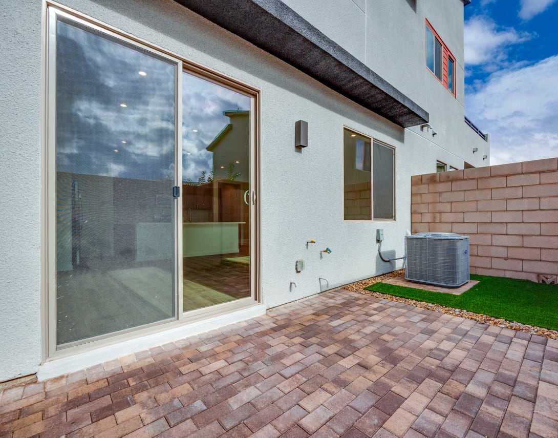 A modern, single-story building with large glass sliding doors leading to a paved patio area with artificial grass, surrounded by a clear blue sky.