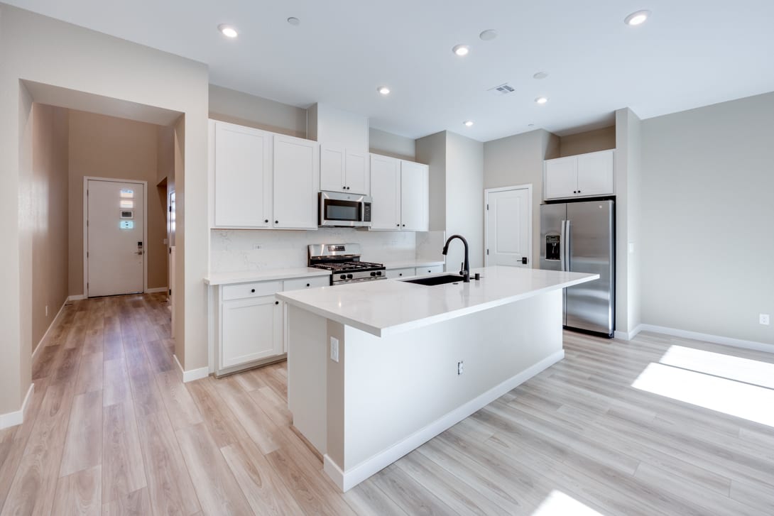 A modern, open-concept kitchen with white cabinets, stainless steel appliances, and a central island with a white countertop, set against a hardwood floor and a hallway leading to another room.
