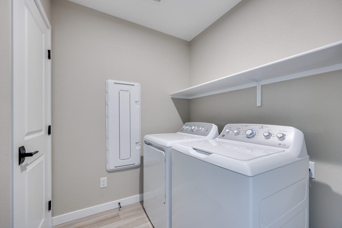 A modern, well-lit laundry room with a white washer and dryer set against a neutral-colored wall, and a small door or closet visible in the background.