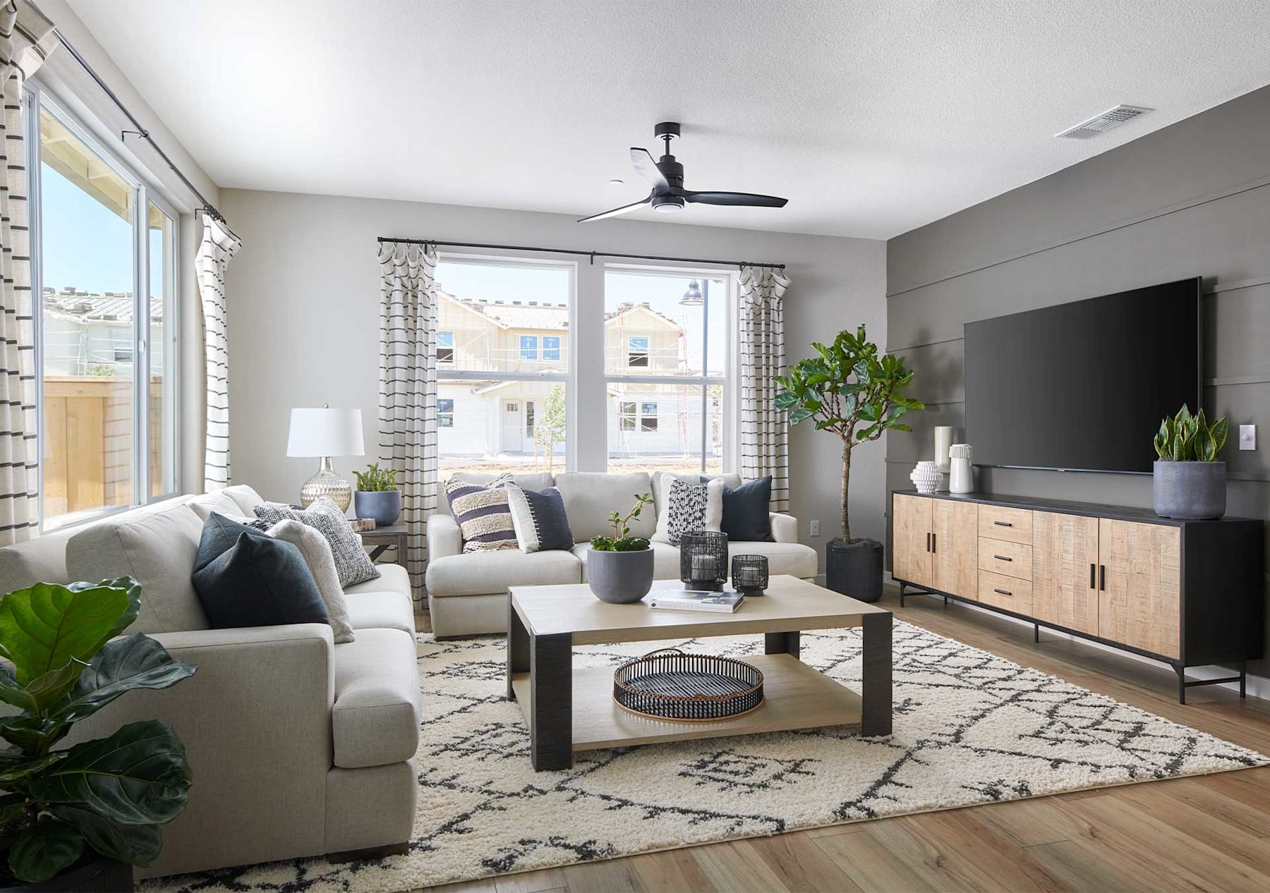 A cozy and modern living room with a gray sofa, a wooden coffee table, and a large TV stand, surrounded by large windows and decorated with potted plants.