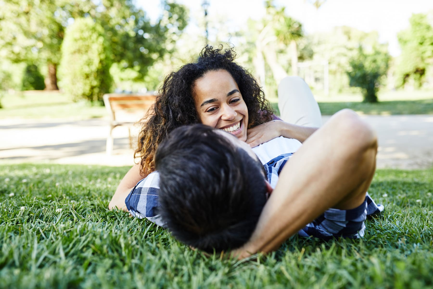 A smiling woman with long, dark hair is lying on the grass in a grassy, tree-lined park setting.