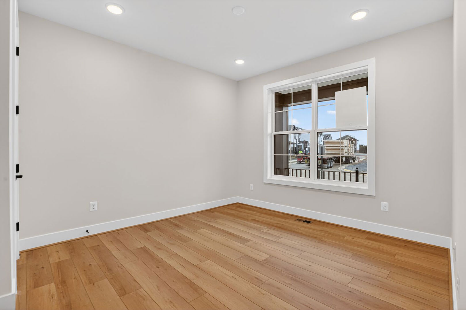 A spacious, well-lit room with hardwood floors and a large window overlooking a residential neighborhood.