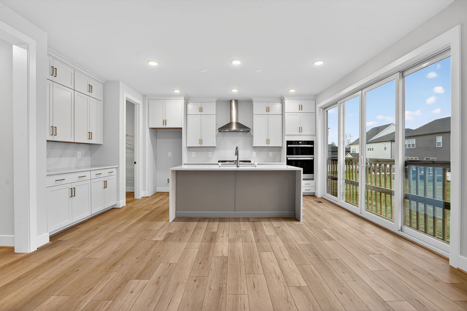 A modern, bright kitchen with white cabinets, a gray island, and large windows overlooking a backyard.