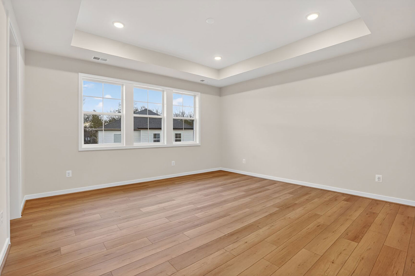 A spacious, well-lit room with hardwood floors, recessed lighting, and large windows overlooking a residential neighborhood.