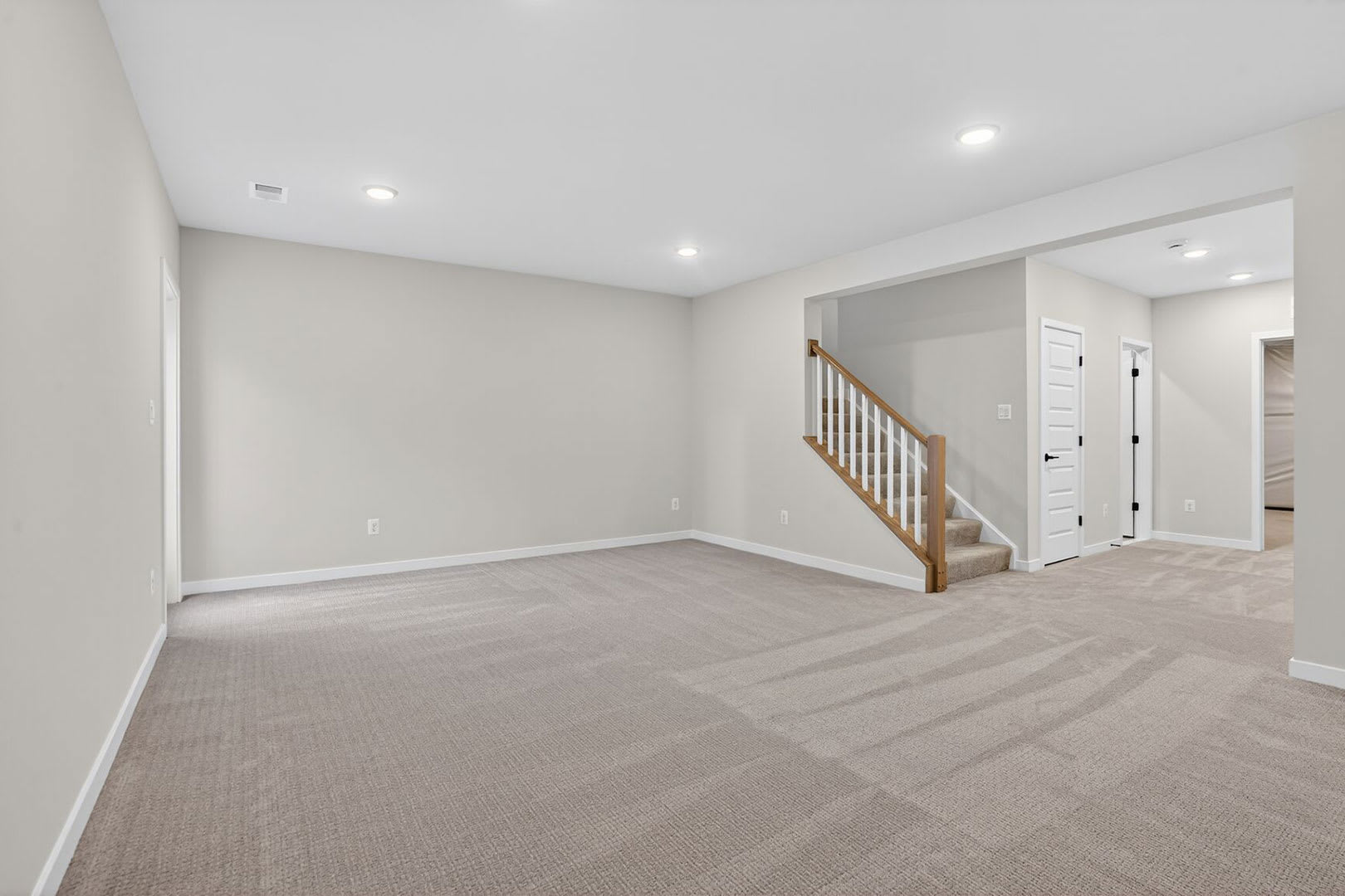 An empty, spacious room with light gray walls, a carpeted floor, and a wooden staircase leading to an upper level. The room appears to be a newly constructed or recently renovated space.