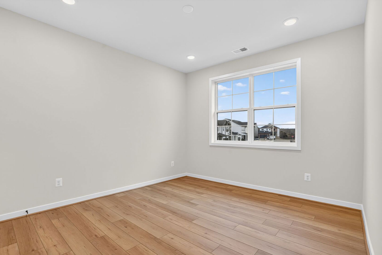 A spacious, well-lit room with a large window overlooking a residential neighborhood, featuring hardwood floors and a neutral color scheme.