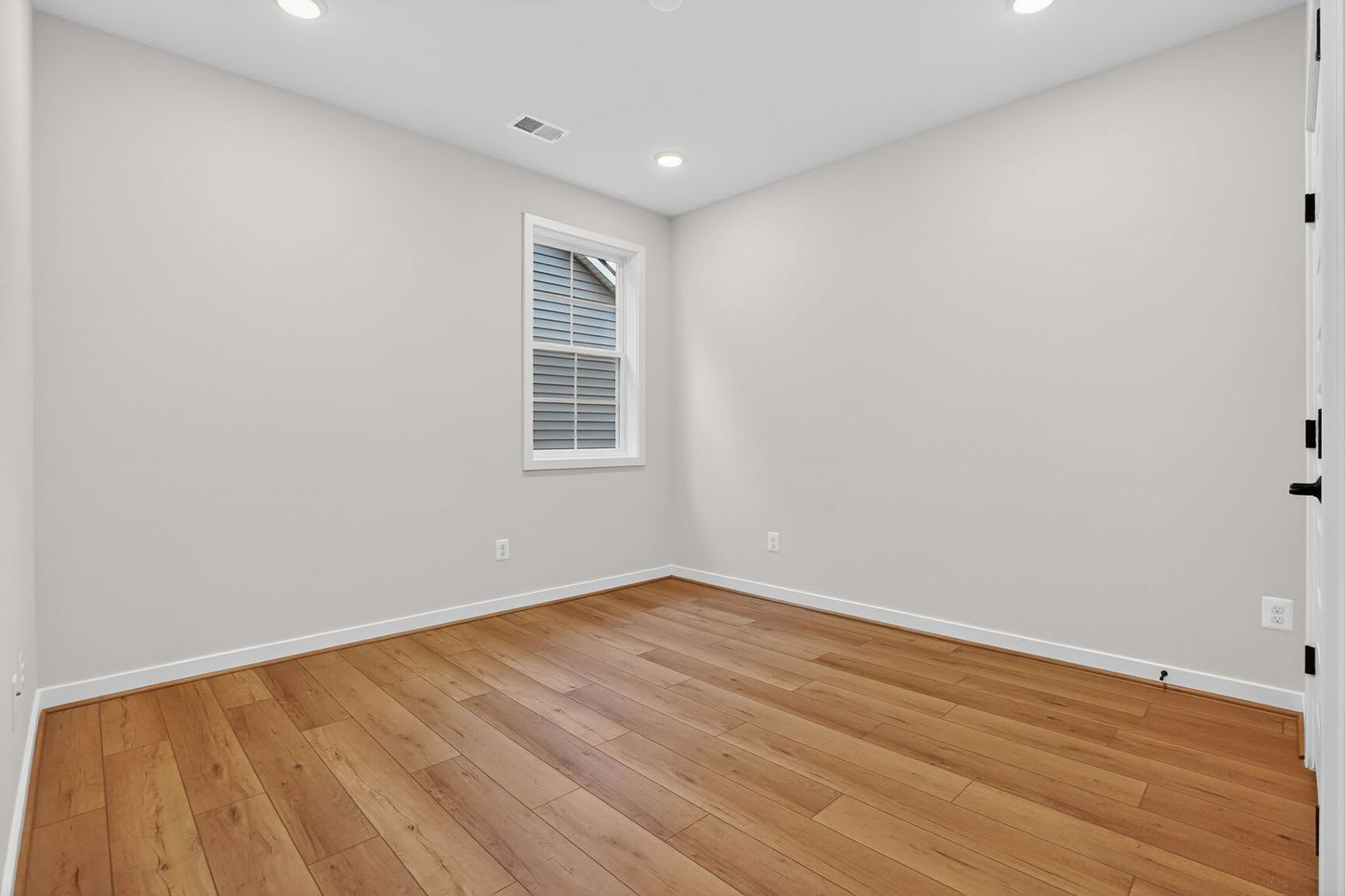 A spacious, well-lit room with light-colored walls and a hardwood floor, featuring a single window and recessed lighting.
