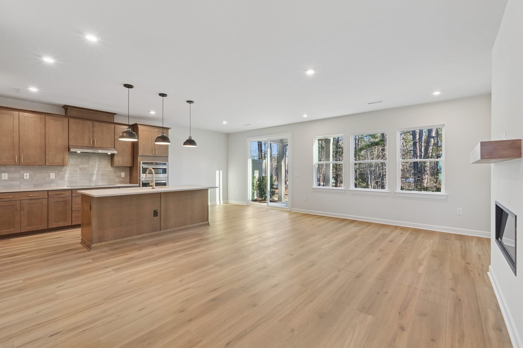 A spacious and modern kitchen with wooden cabinets, a white countertop, and large windows overlooking a wooded area.