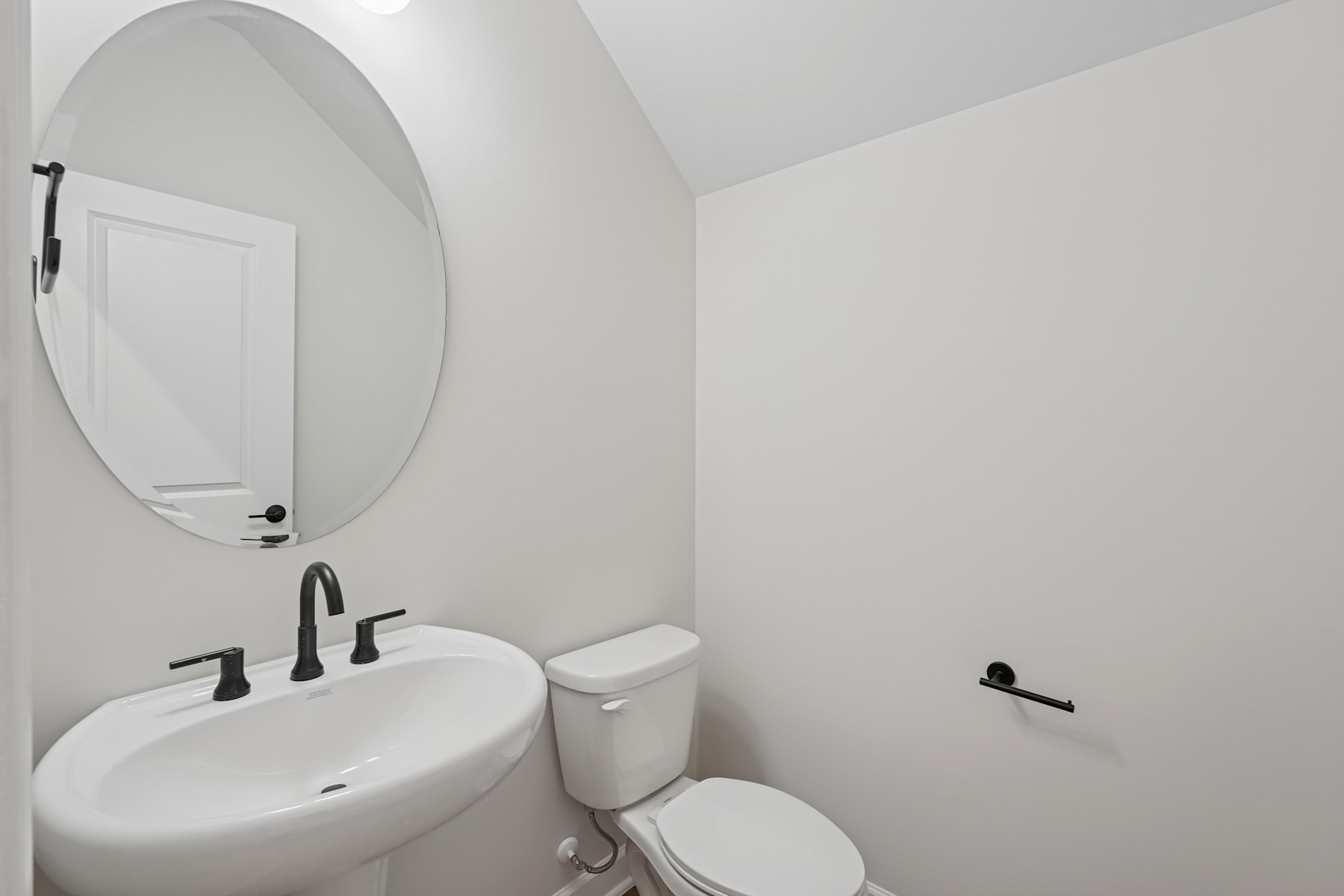 A modern, minimalist bathroom with a white oval mirror, a white sink, and a toilet against a plain white wall.