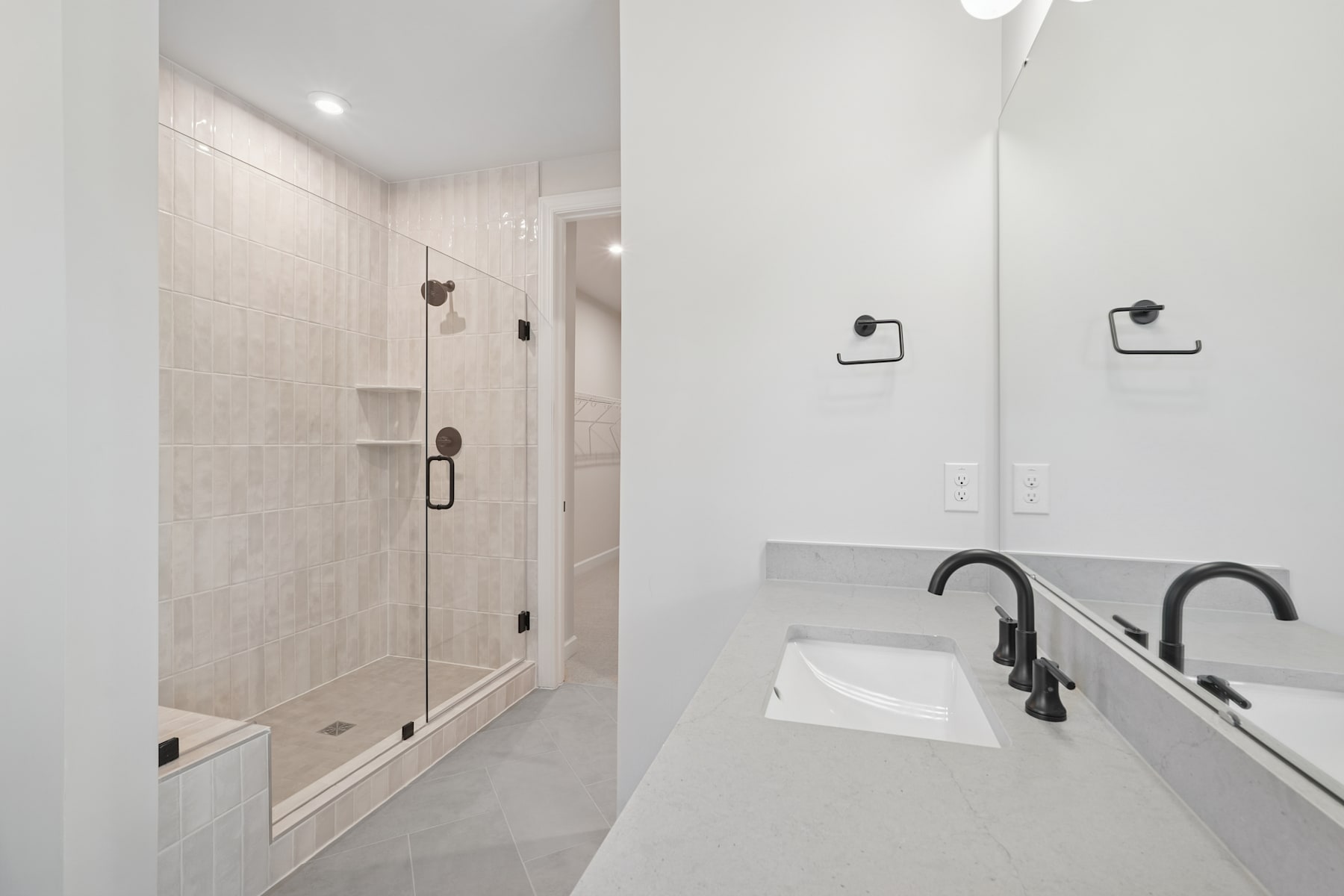 A modern, minimalist bathroom with a glass shower enclosure, a white vanity with a sink, and black fixtures against a light-colored tiled wall.