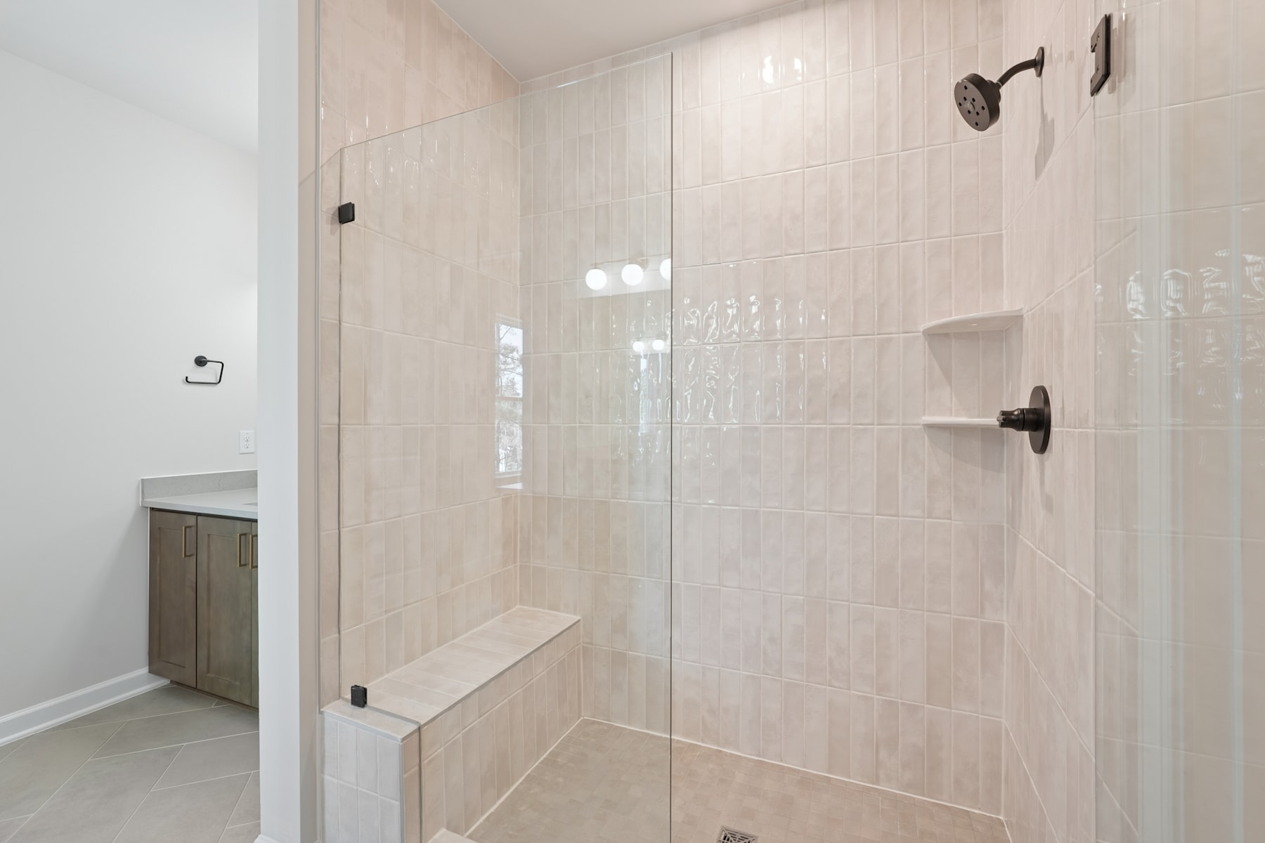 A modern, minimalist bathroom with a glass shower enclosure, beige tile walls, and a built-in bench.