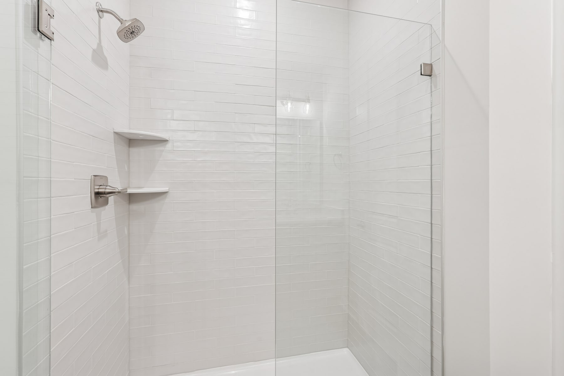 A modern, minimalist bathroom with a glass shower enclosure and white tiled walls, featuring a showerhead and shelves for storage.