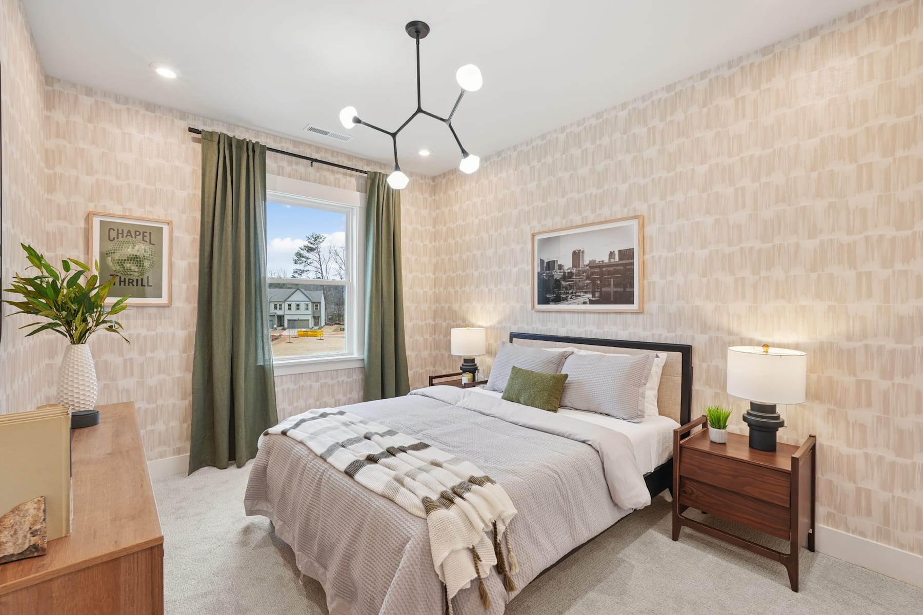 A cozy and well-decorated bedroom with a large bed, wooden nightstands, and a modern chandelier overhead, set against a backdrop of patterned wallpaper and a window overlooking an outdoor scene.