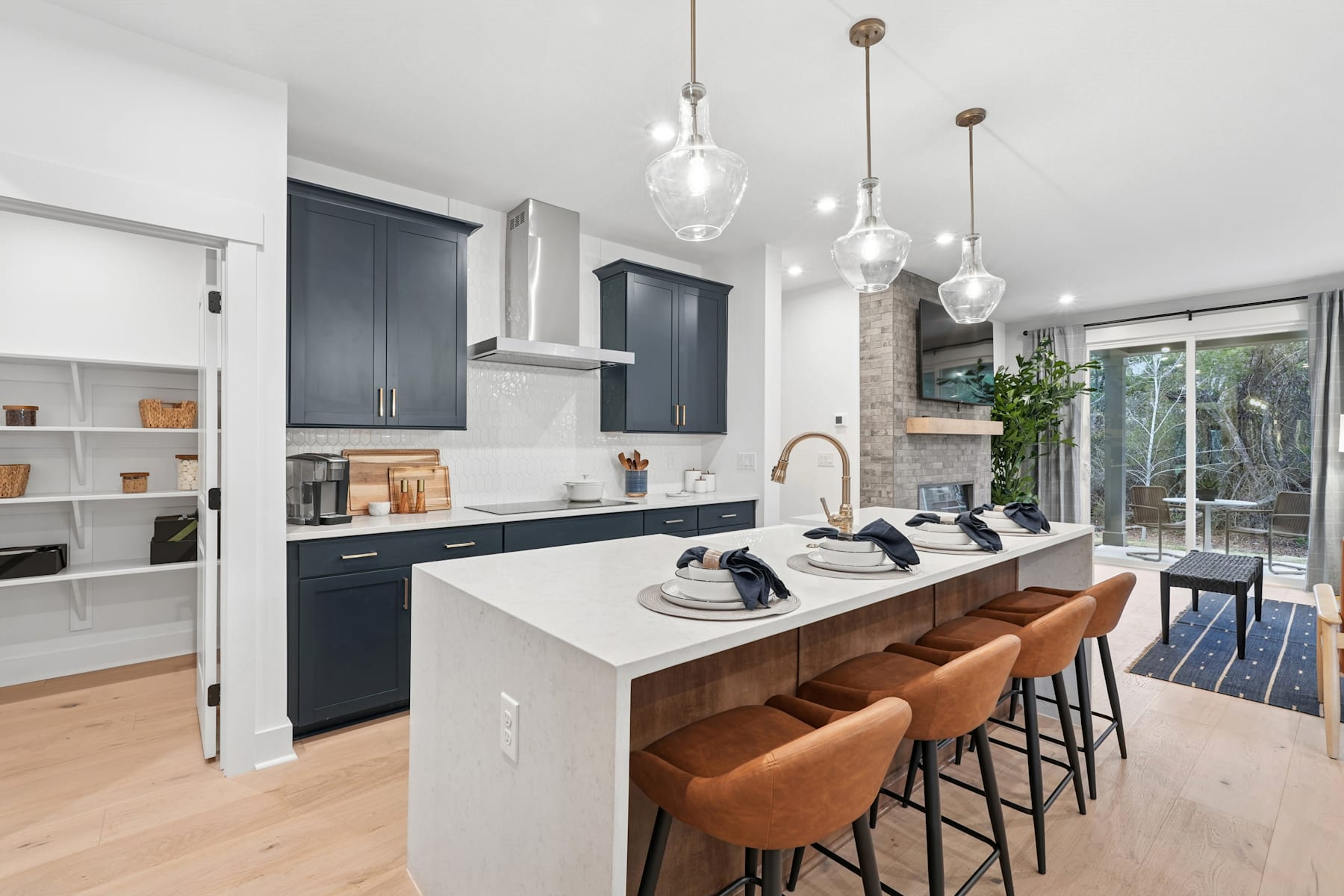 A modern and stylish kitchen with dark cabinets, white countertops, and pendant lights, featuring a breakfast bar with orange stools and a view of the outdoor area through a large window.