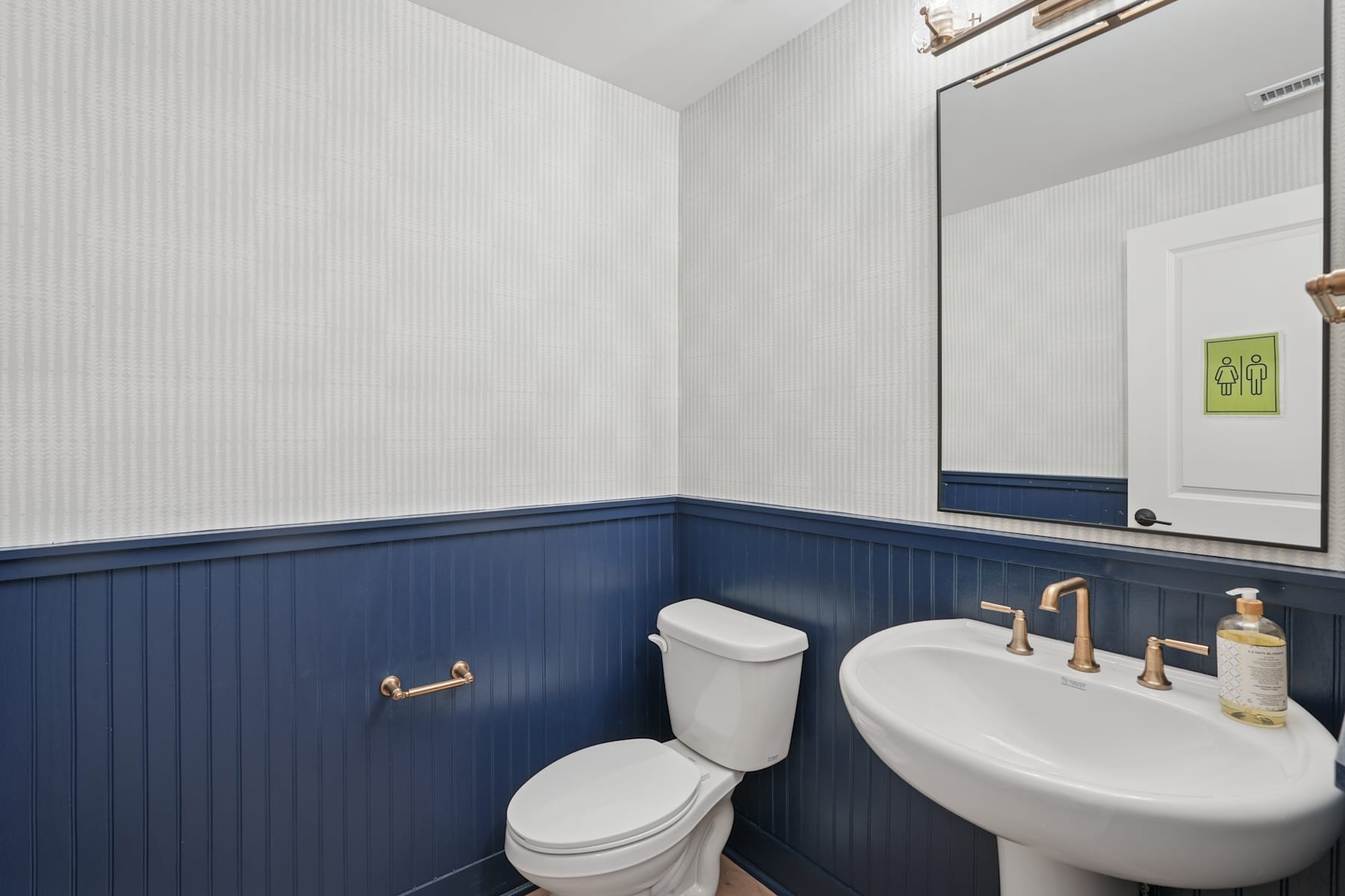 A modern bathroom with white walls, a navy blue wainscoting, a white toilet, and a white sink with gold fixtures.