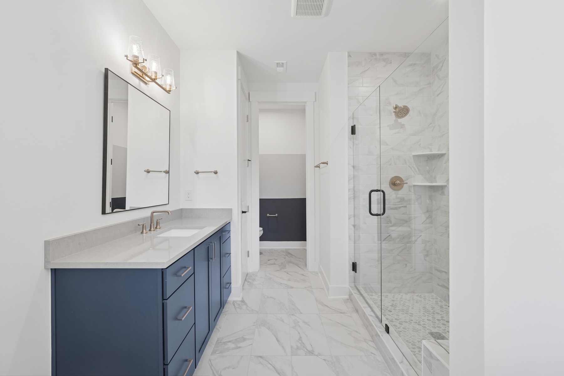 A modern, minimalist bathroom with a navy blue vanity, a large mirror, and a marble-tiled floor leading to a shower area.