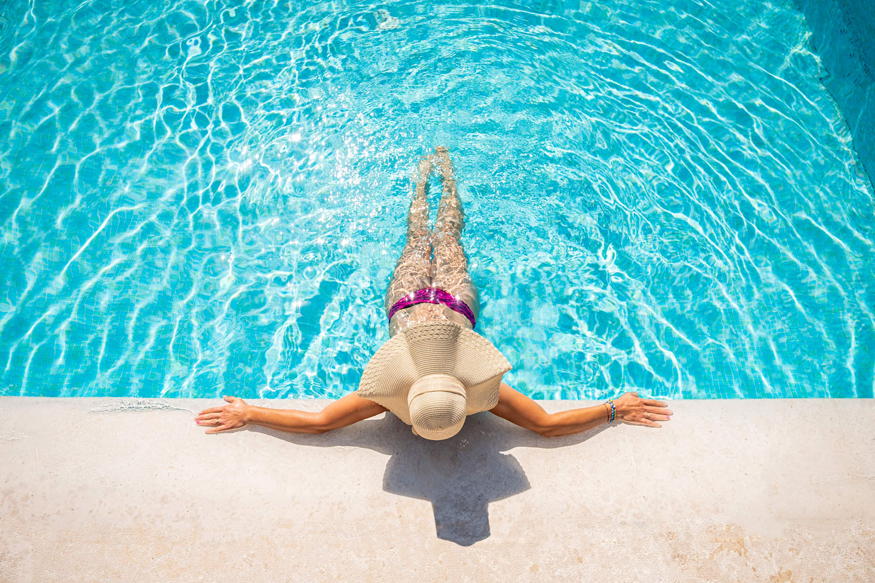 A person in a pink swimsuit is relaxing on the edge of a clear, turquoise swimming pool, with their legs dangling in the water.