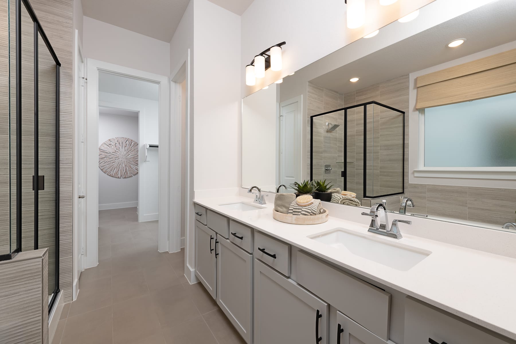 A modern and well-designed bathroom with a double vanity, white countertops, and a large mirror with a decorative frame. The room has a clean and minimalist aesthetic, with recessed lighting and a neutral color palette.