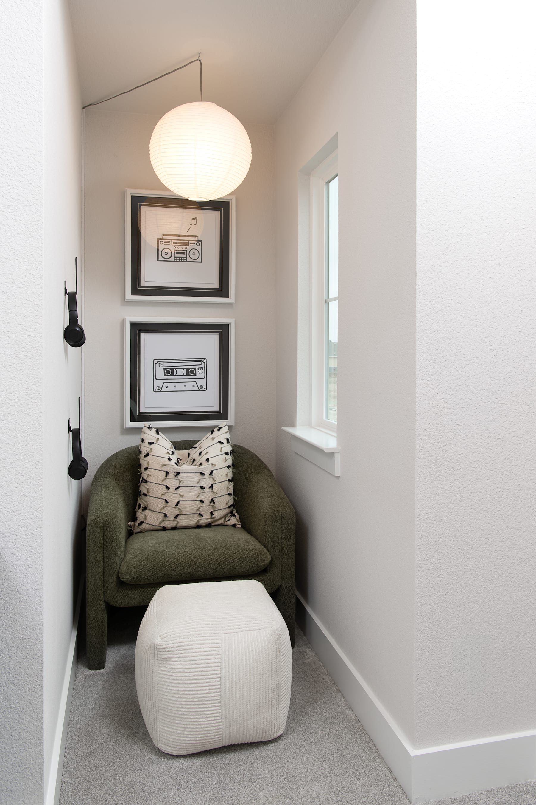 A cozy and minimalist reading nook with a green armchair, a white pouf, and framed artwork on the walls, illuminated by a round pendant light.