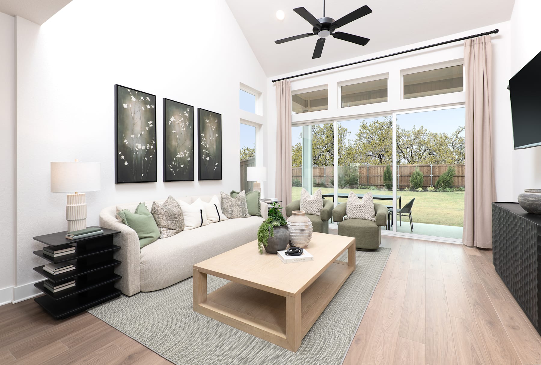 A modern and minimalist living room with large windows overlooking a lush outdoor area, featuring a sleek black ceiling fan, abstract artwork, and a wooden coffee table.