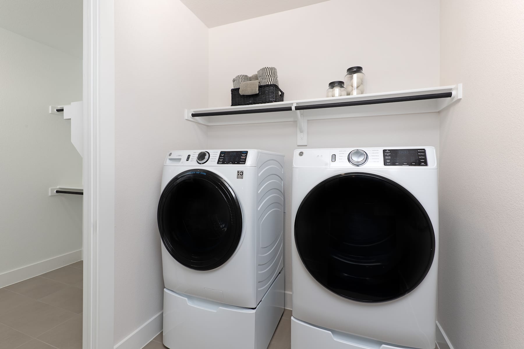 Two modern white washing machines and dryers are placed side by side in a clean, minimalist laundry room setting with a white wall and a shelf above the appliances.