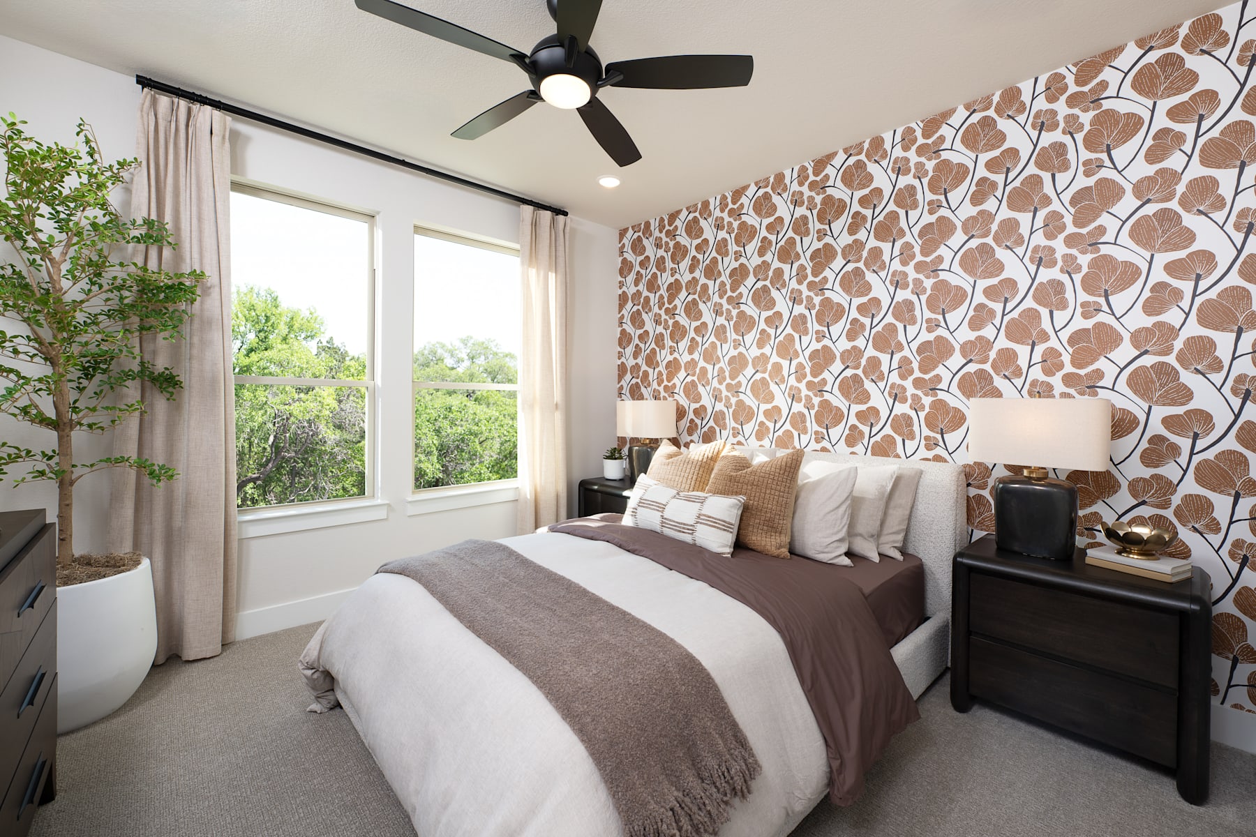 A cozy bedroom with a patterned accent wall, a ceiling fan, and a large window overlooking greenery outside.