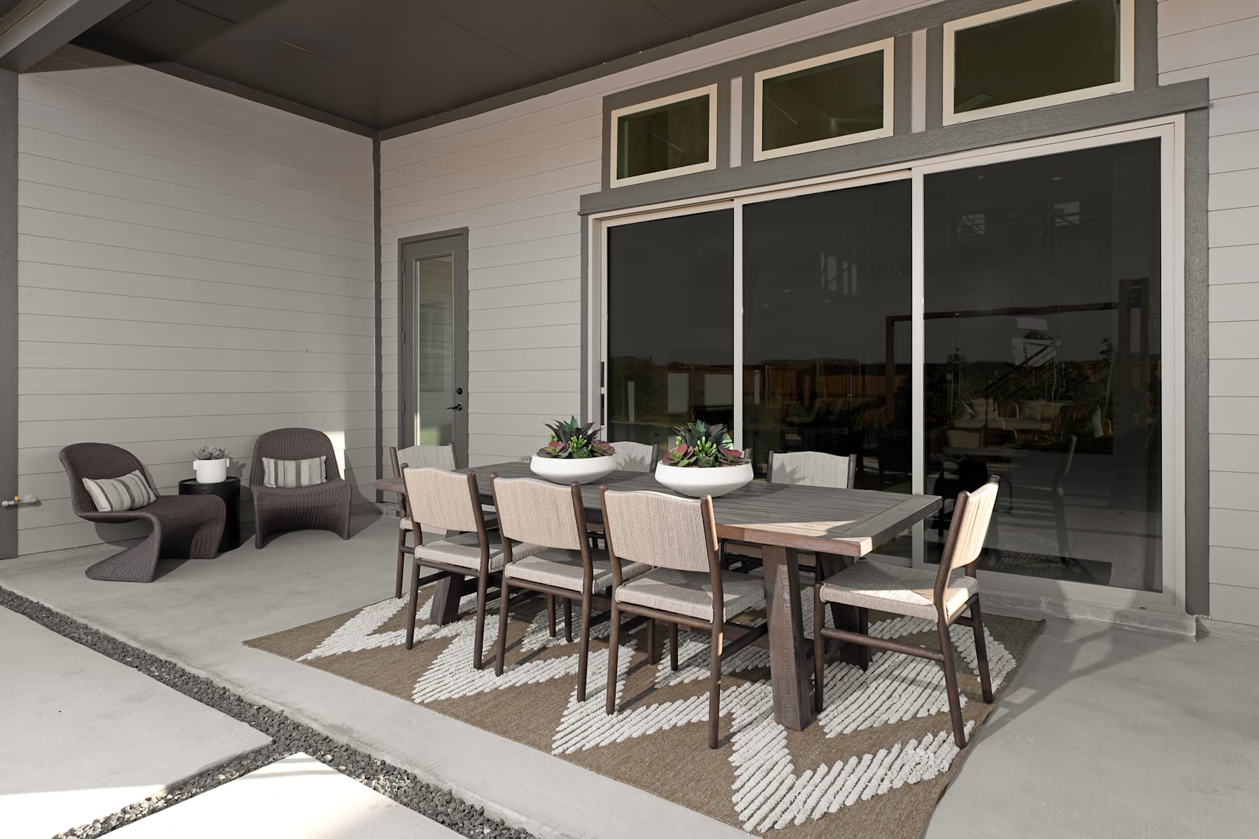A modern and minimalist dining area with a wooden table, chairs, and a patterned rug, surrounded by large windows that provide a view of the exterior.