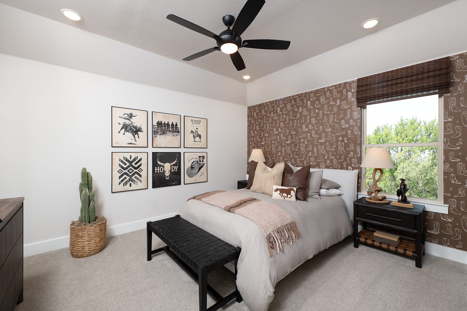 A cozy and well-decorated bedroom with a ceiling fan, framed artwork on the walls, and a large window overlooking greenery outside.