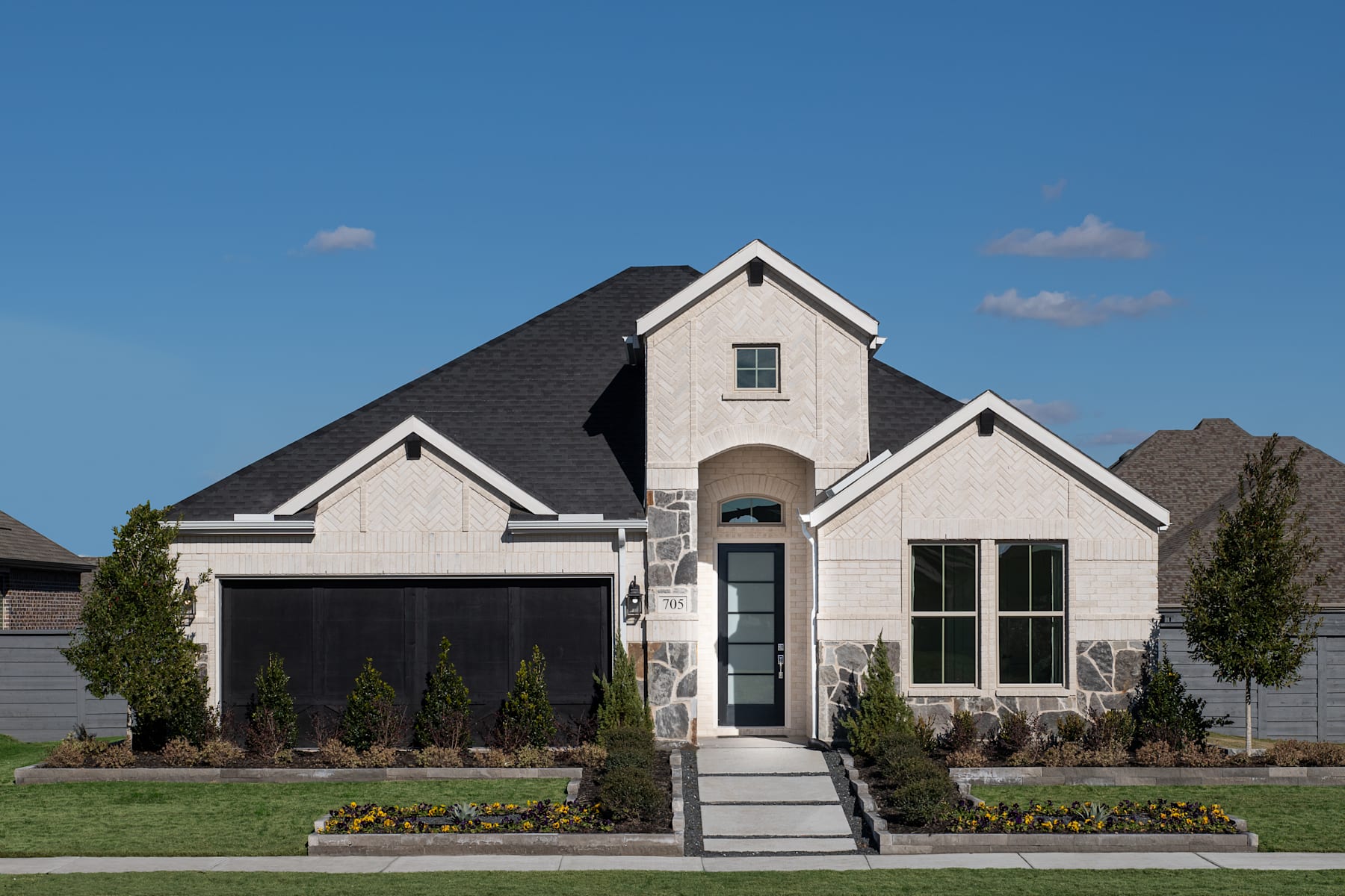 A two-story house with a gabled roof, stone and siding exterior, and a landscaped front yard with shrubs and flowers, set against a clear blue sky with a few clouds.