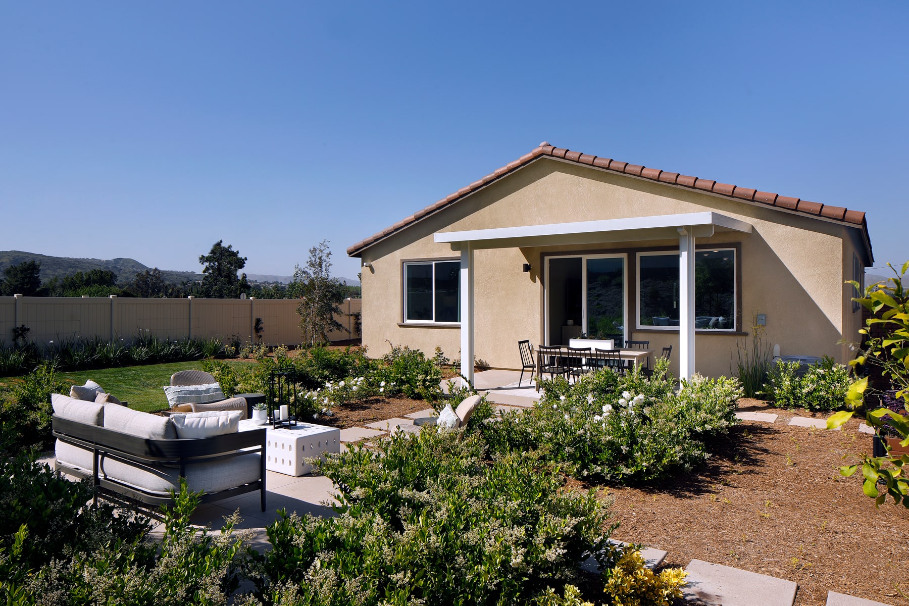 A cozy, single-story house with a well-maintained garden and patio area in the foreground, set against a backdrop of mountains and a clear blue sky.