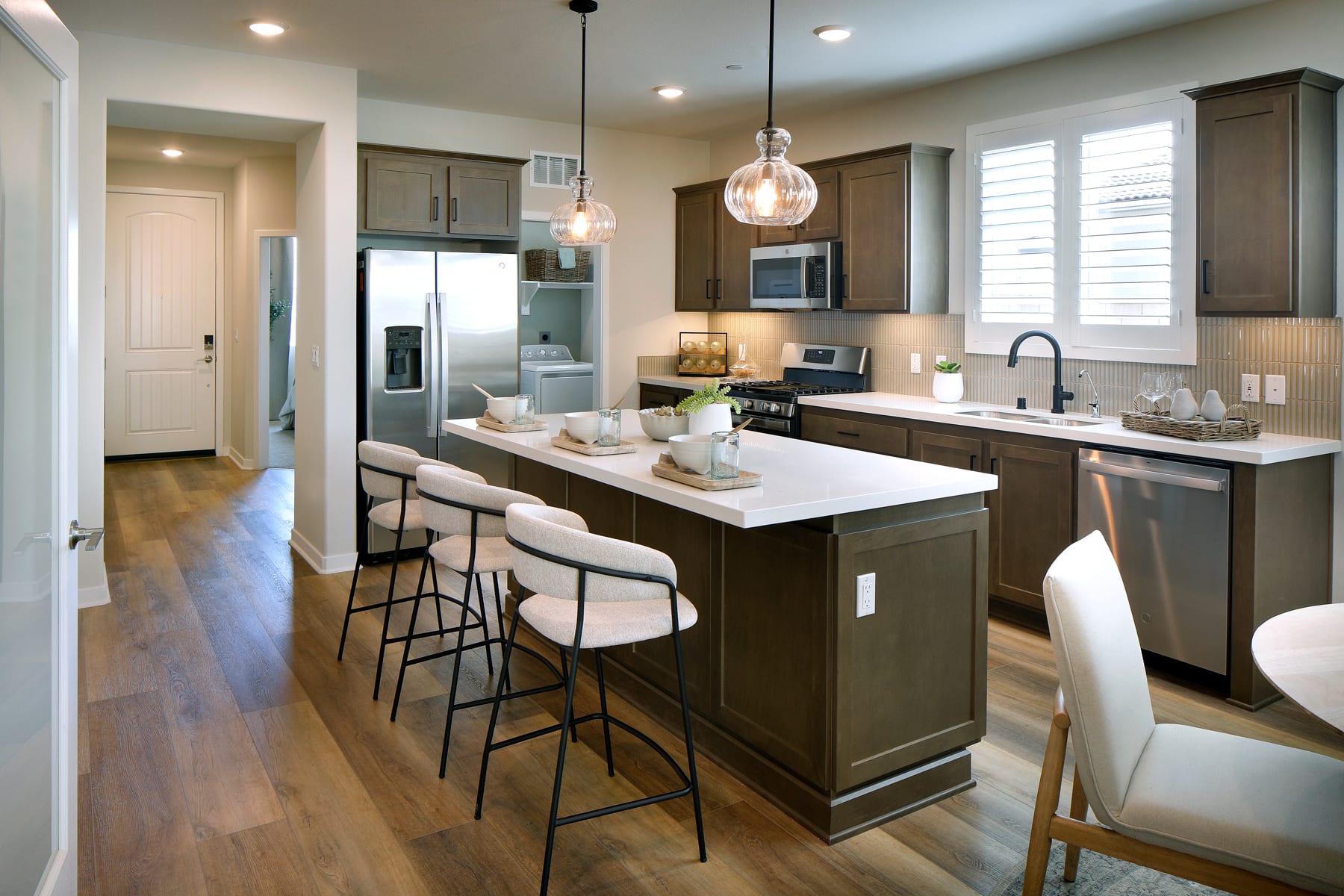 A modern and well-designed kitchen with dark wood cabinets, a large island with seating, and pendant lighting fixtures, set against a bright and airy background with hardwood floors and a view of an adjacent room.