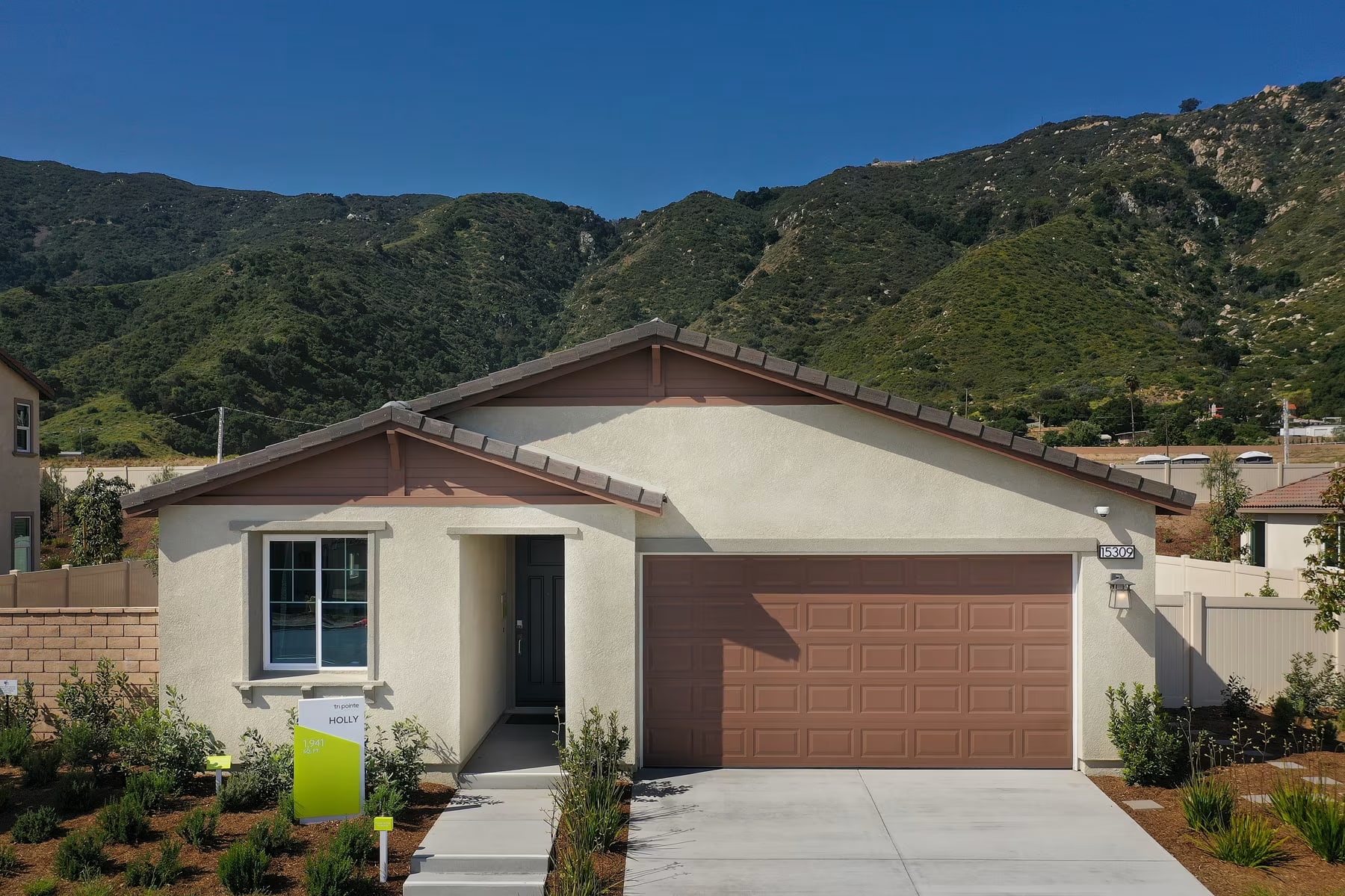 A single-story residential house with a garage door and a small yard in the foreground, surrounded by lush green hills and mountains in the background.